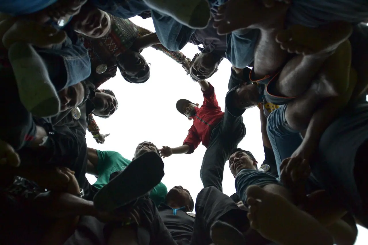 Looking up from the ground at a circle of participants leaning in together with their hands reaching toward the centre against a bright sky.