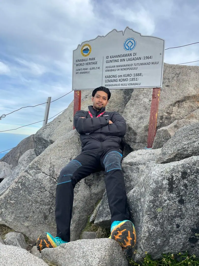 Azidzul sitting with arms crossed on granite boulders beneath the Kinabalu Park World Heritage summit sign at Low’s Peak, 4,095.2 metres.