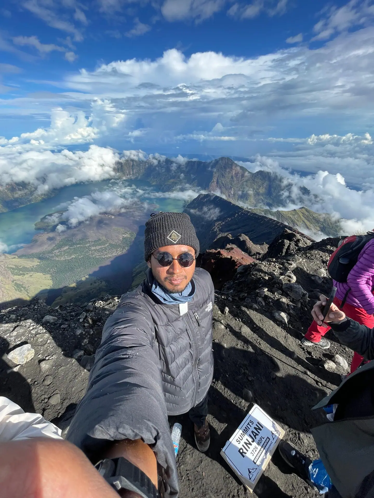 Azidzul in a beanie and round sunglasses on the Mount Rinjani summit ridge, above the crater lake and a sea of cloud, next to the 3,726 metre summit sign.