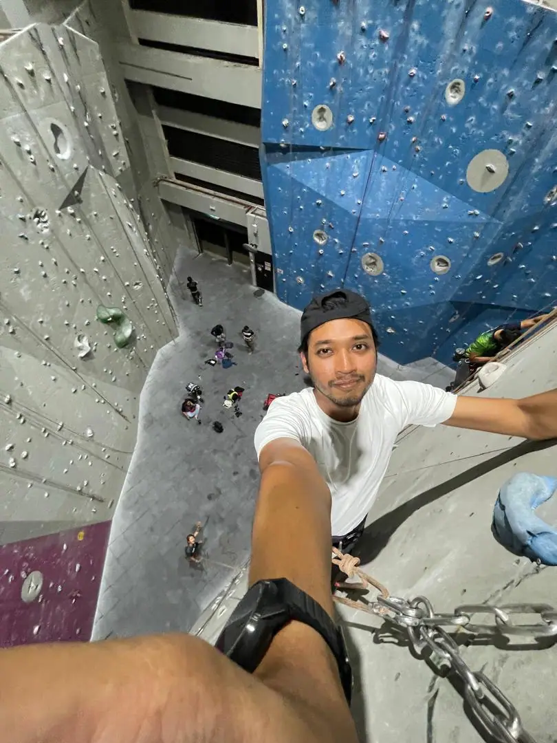Azidzul smiling at the camera while clinging to an indoor climbing wall, with the floor and other climbers far below.
